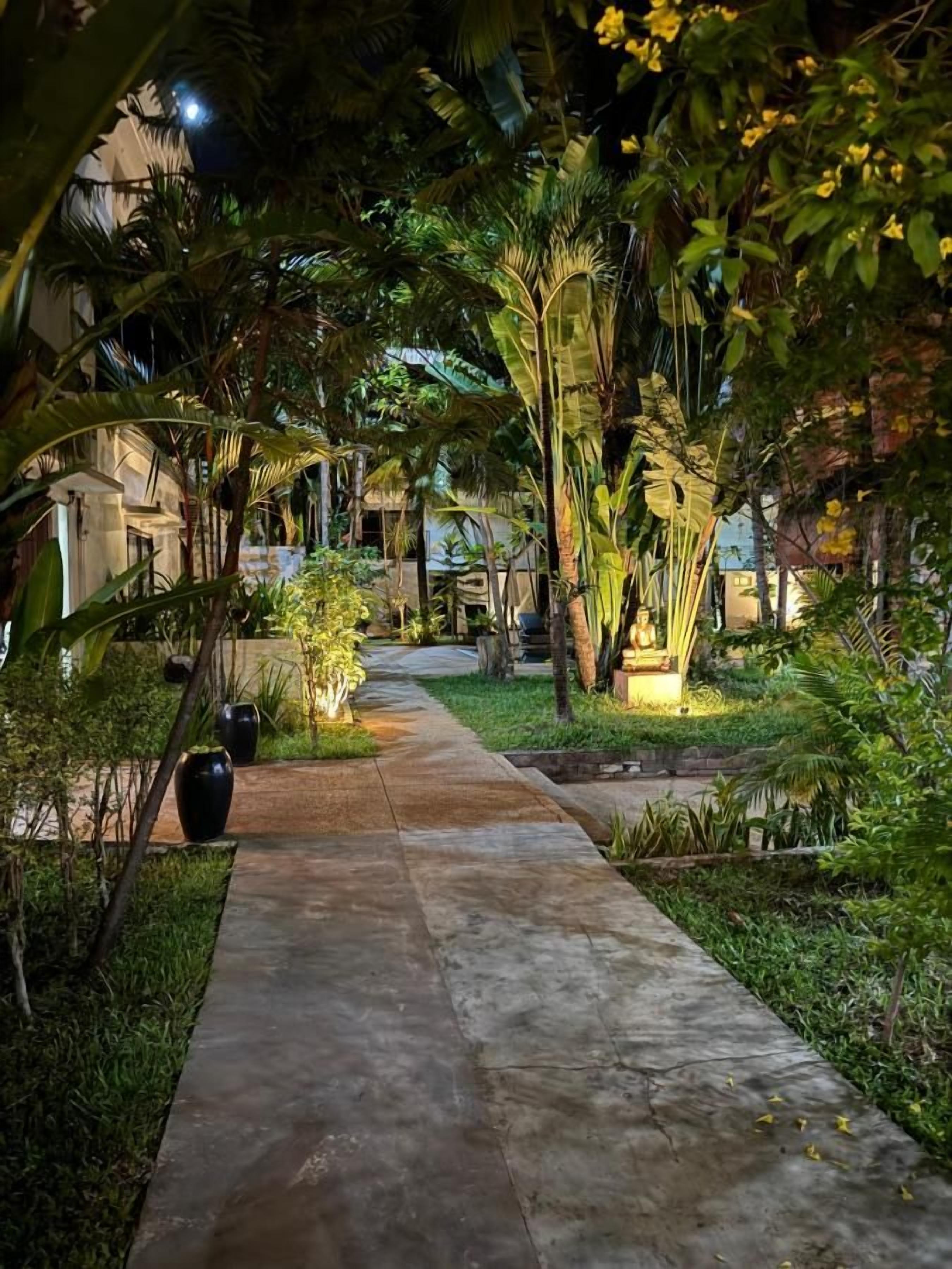 A pathway flanked by lush greenery and palm trees, leading towards a building.