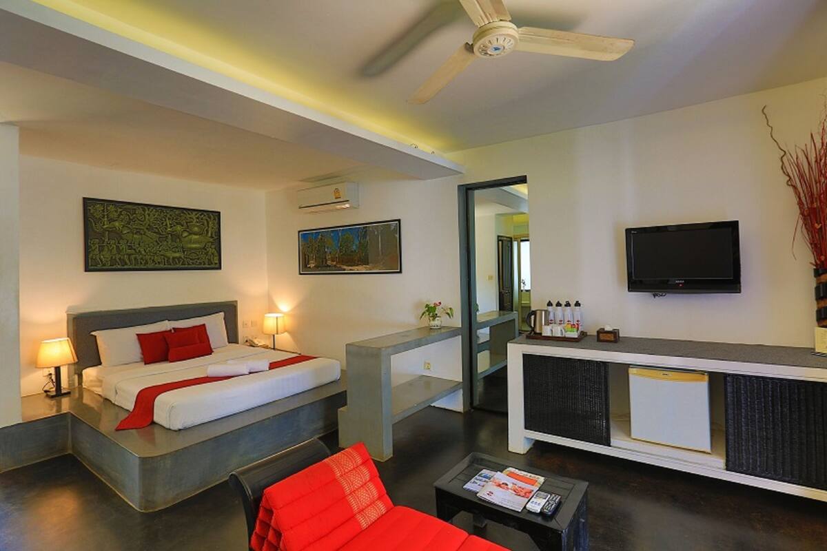 A modern hotel room with a bed, a seating area with a red chair, a coffee table, a flat-screen TV, and a small refrigerator.
