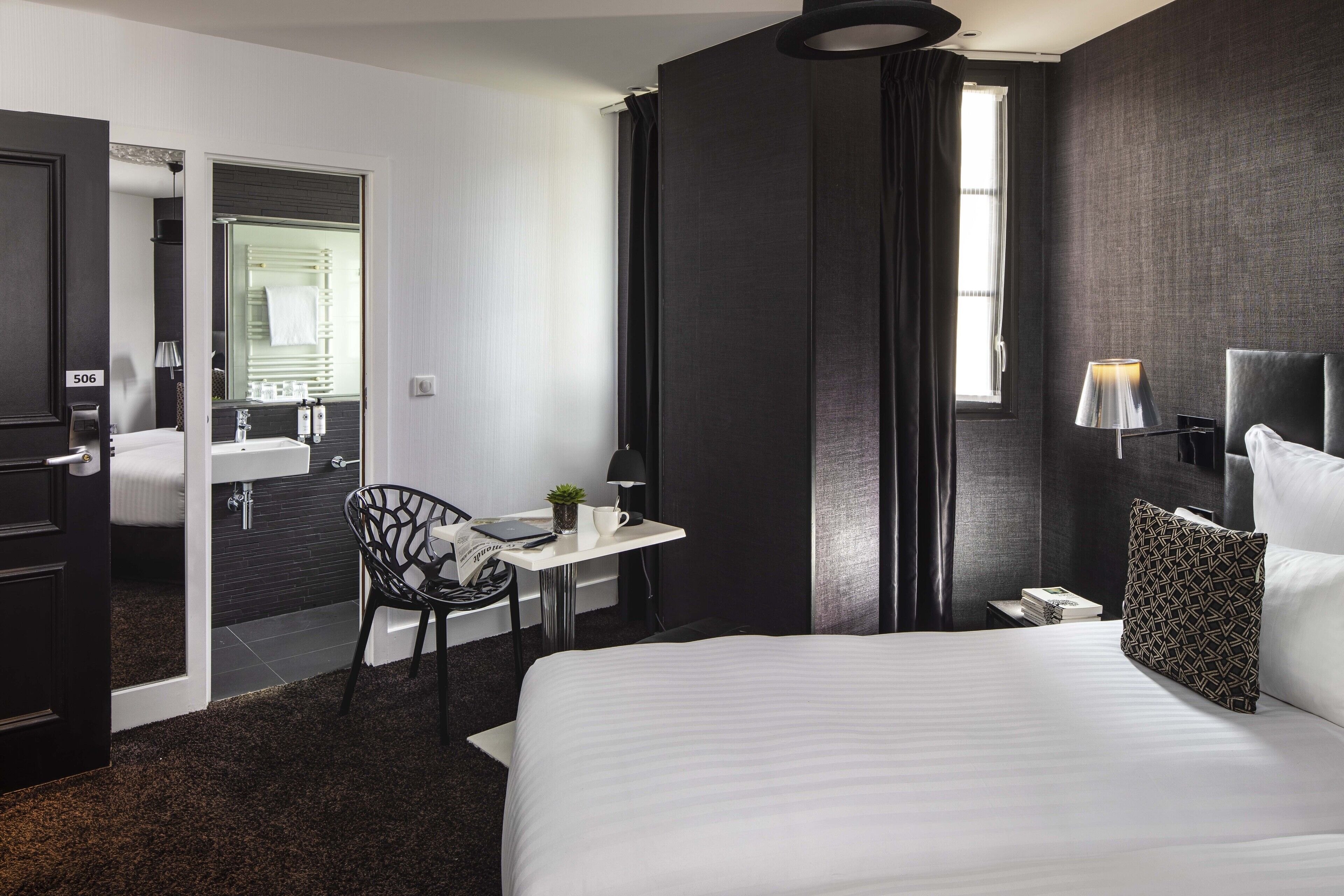 Ein Hotelzimmer mit einem großen Bett, einem Schreibtisch mit Stuhl, einem kleinen Tisch mit Pflanze, einem Nachttisch mit Lampe und einem Fenster mit Vorhängen.