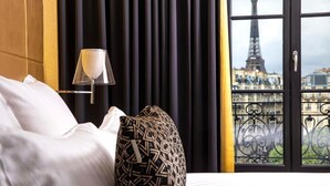 Une chambre d’hôtel avec un lit, un coussin à motifs, une lampe et une vue sur la Tour Eiffel, visible à travers une fenêtre ornée de barreaux en fer.