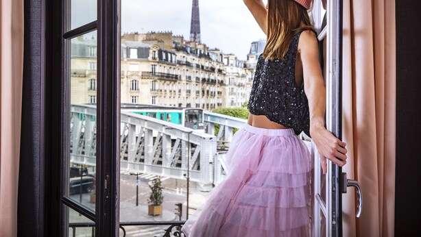 Une personne appuyée contre un rebord de fenêtre, d’où elle contemple un paysage urbain avec des bâtiments historiques et, au loin, la Tour Eiffel.
