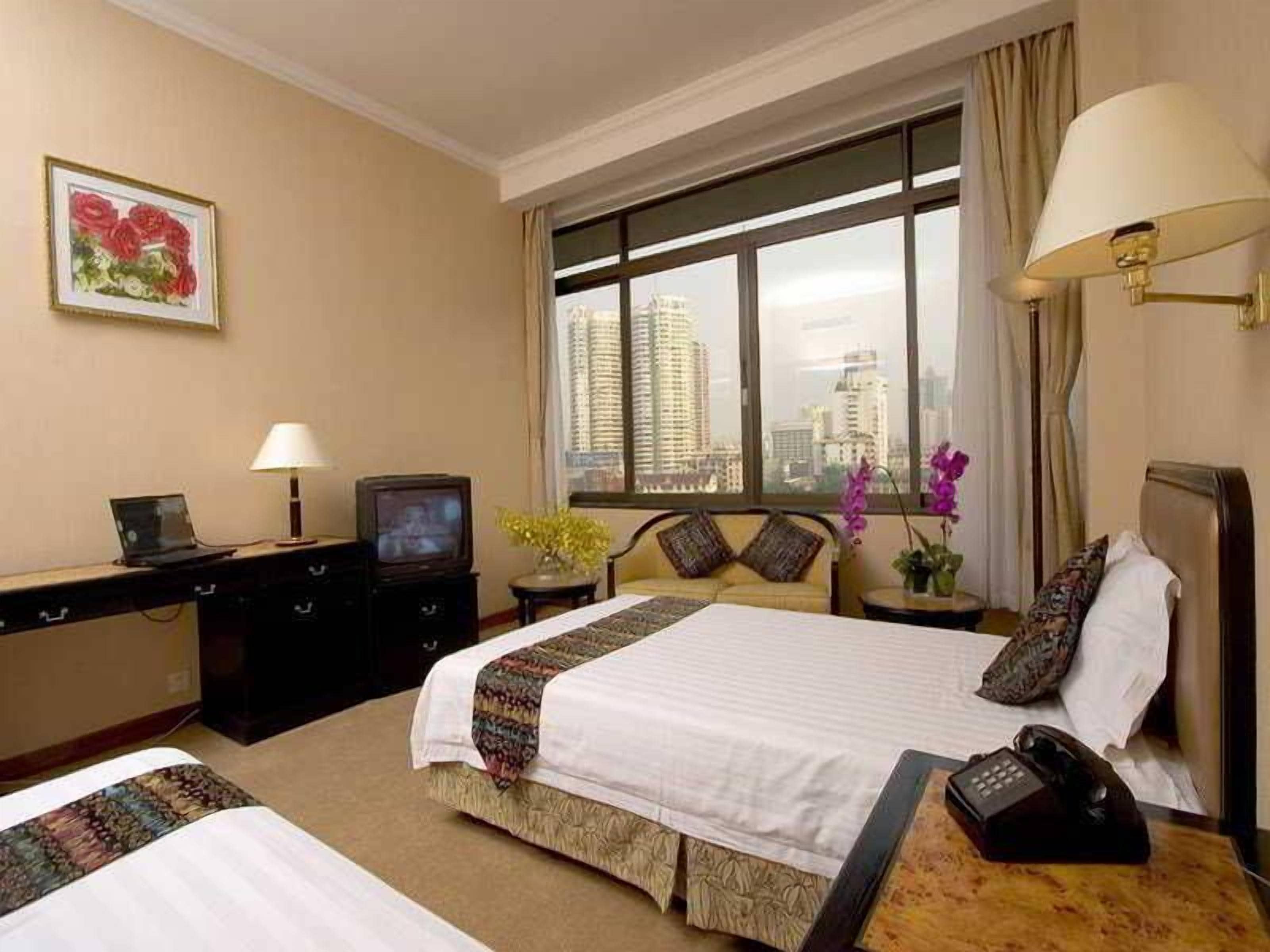 A hotel room with a bed, bedside table, lamp, TV, and a view of the city.