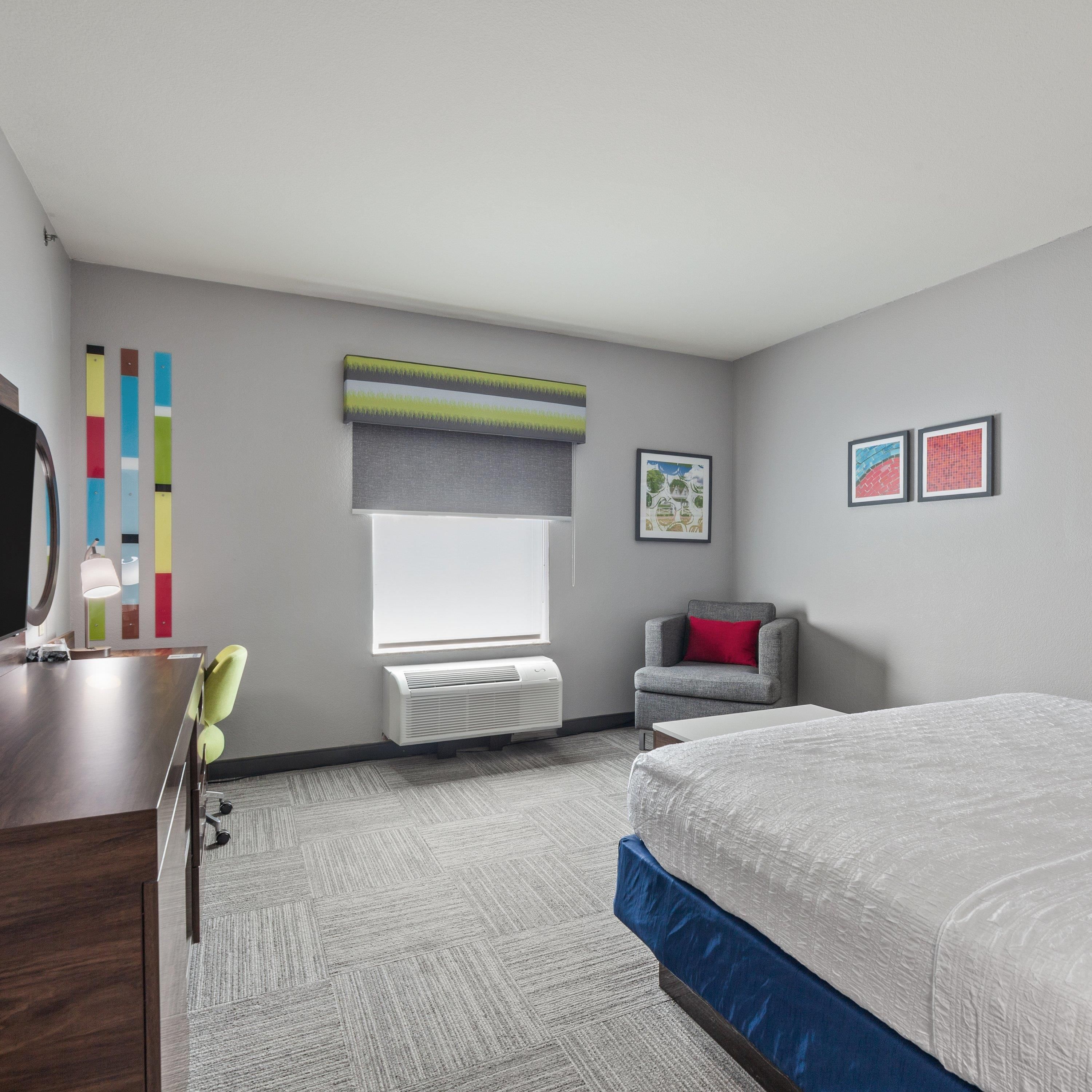 Une chambre d’hôtel comprenant un lit, un bureau, une chaise, une fenêtre avec des stores, un radiateur et deux tableaux encadrés accrochés au mur.