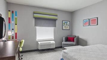Une chambre d’hôtel comprenant un lit, un bureau, une chaise, une fenêtre avec des stores, un radiateur et deux tableaux encadrés accrochés au mur.