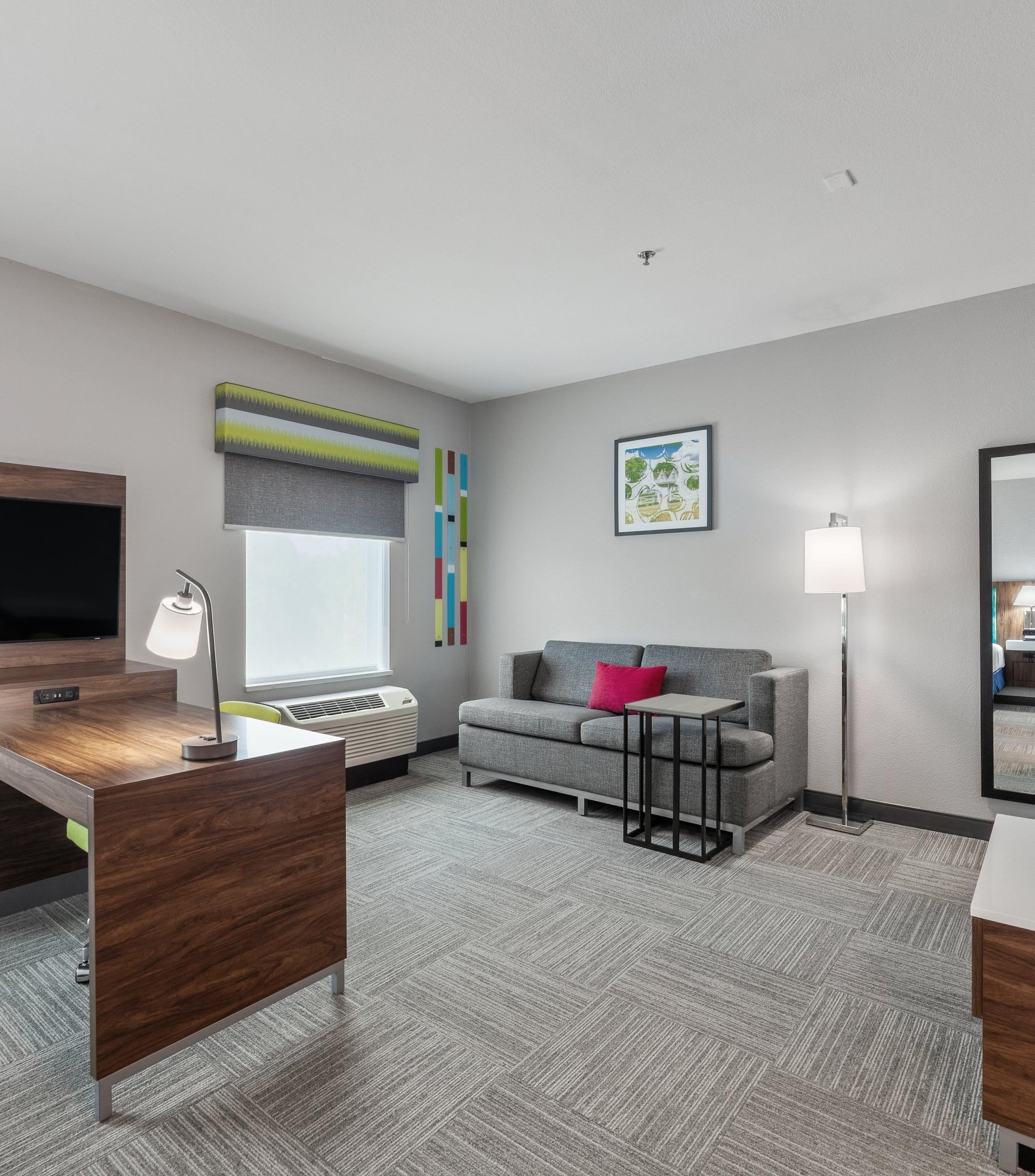 Quarto de hotel moderno com televisão de tela plana, escrivaninha com luminária, sofá cinza com almofada vermelha e uma grande janela com vista para árvores.