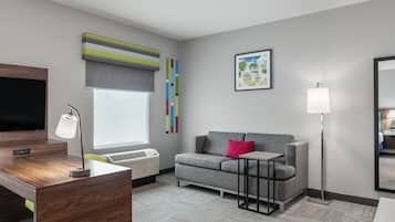Quarto de hotel moderno com televisão de tela plana, escrivaninha com luminária, sofá cinza com almofada vermelha e uma grande janela com vista para árvores.