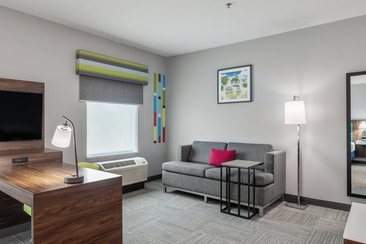 A modern hotel room with a flat-screen TV, a desk with a lamp, a grey sofa with a red pillow, and a large window with a view of trees.