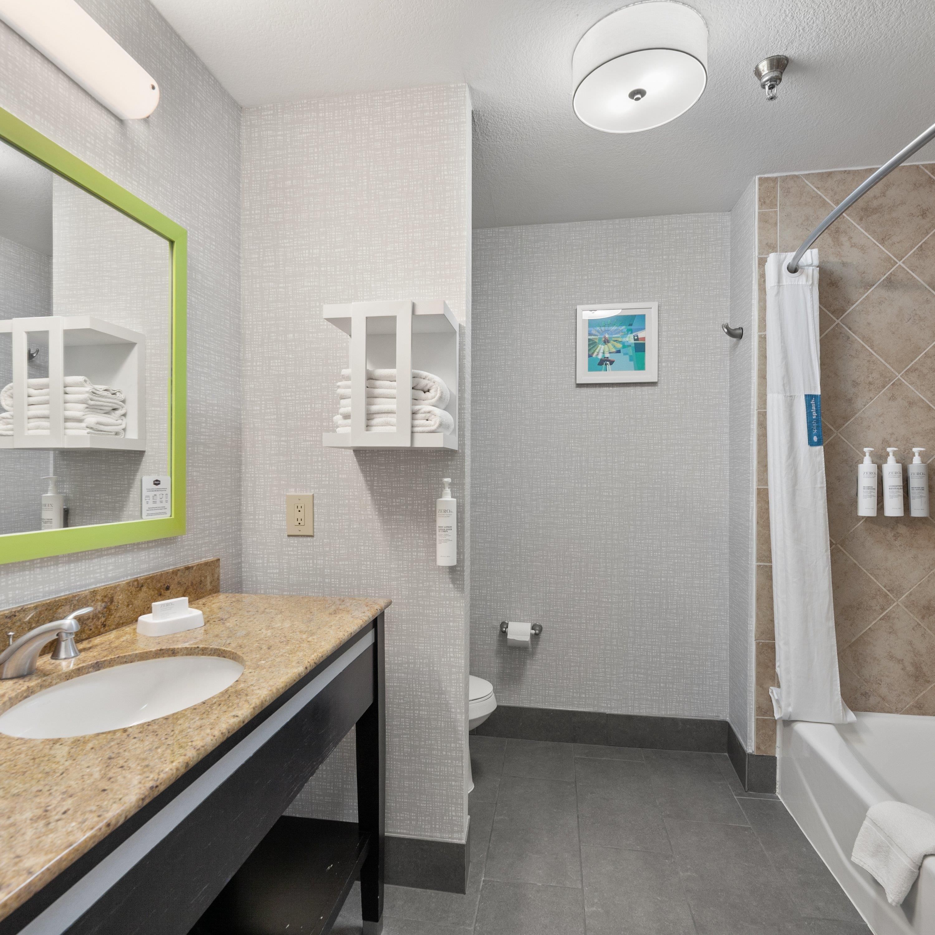 A modern bathroom with a granite countertop sink, a mirror with a green frame, a toilet, a shower with a beige curtain, and a framed picture on the wall.