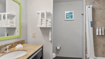 A modern bathroom with a granite countertop sink, a mirror with a green frame, a toilet, a shower with a beige curtain, and a framed picture on the wall.
