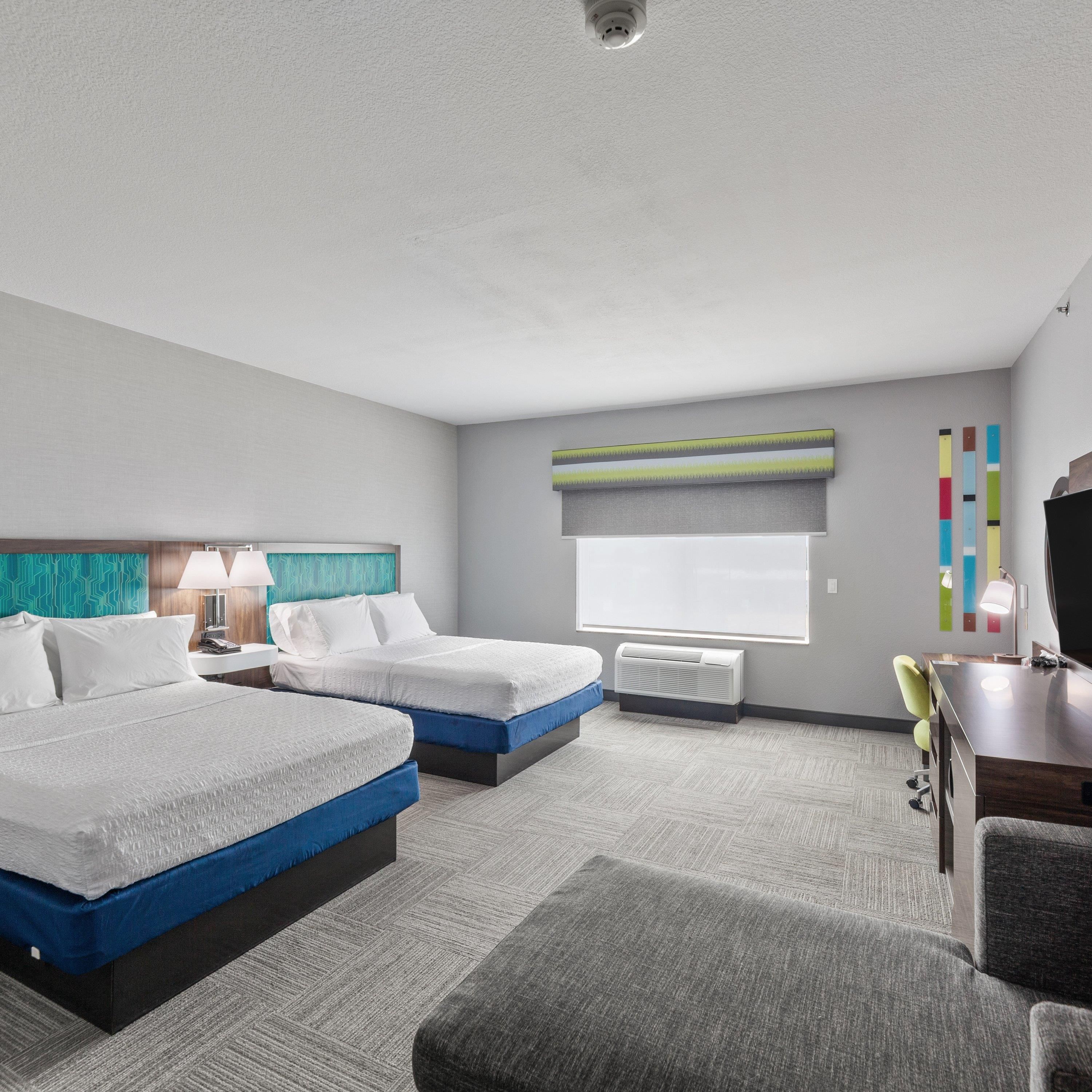 Ein Hotelzimmer mit zwei Betten, einem Schreibtisch, einem Sessel, einem Fernseher und einem Fenster mit Jalousien.