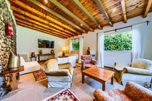 A cozy living room with a stone wall, wooden ceiling beams, a fireplace, and a large window with a view of greenery. - Soft Catedral (San Carlos de Bariloche)