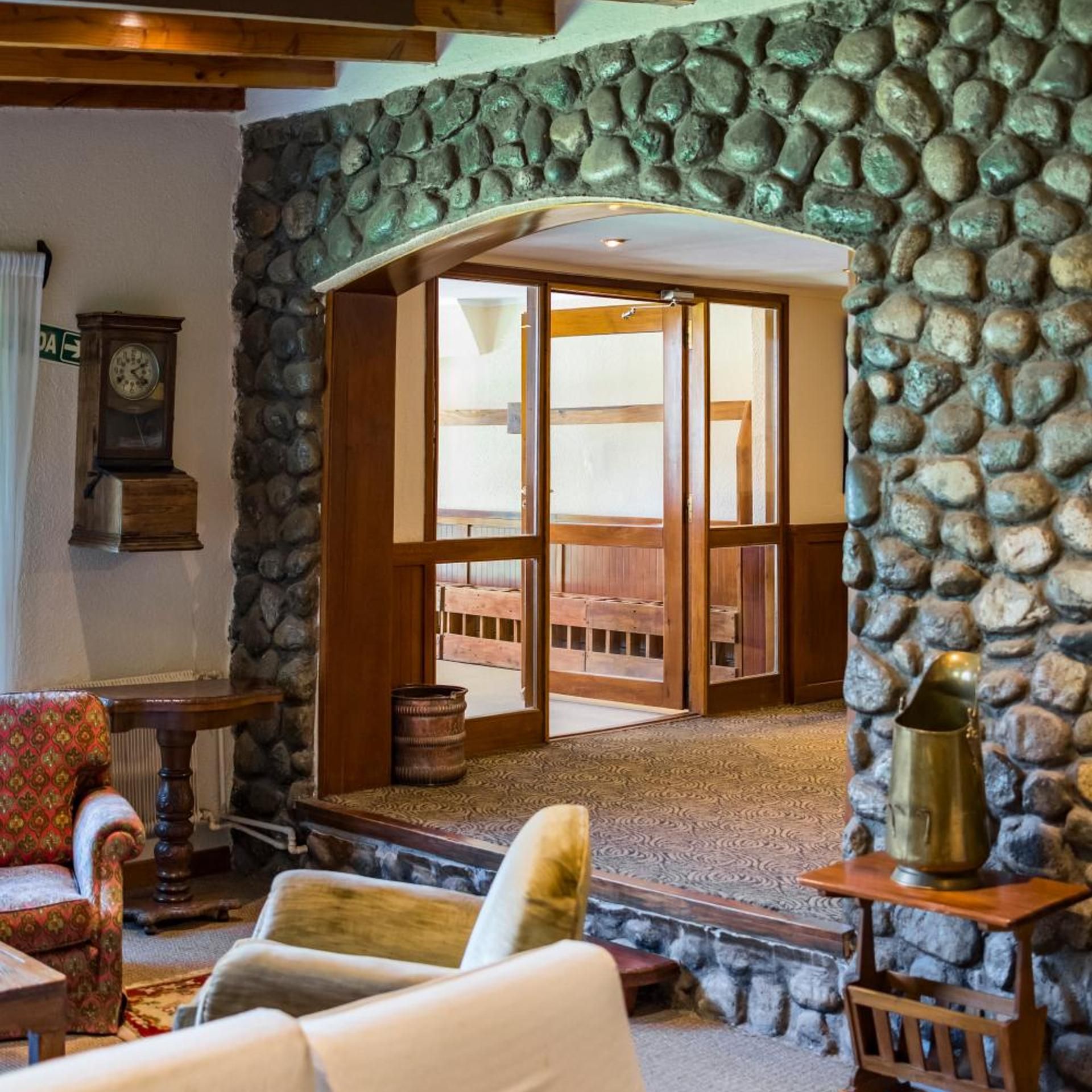 A room with a stone wall, a wooden door leading to an outdoor area, a vintage clock, and a rustic chair.
