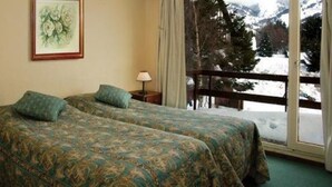 Una habitación con dos camas, un cuadro enmarcado en la pared, una mesita de noche con lámpara y vistas a montañas nevadas a través de un balcón.