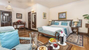 Habitación de hotel con cama, una silla, una mesa con fruta y vino, y una lámpara.