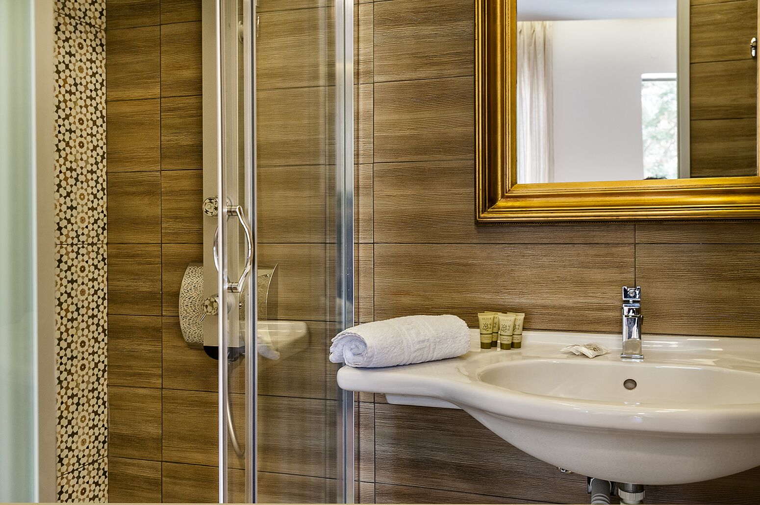 A bathroom with a glass shower enclosure, a white sink with a gold mirror, and patterned tiles on the walls.