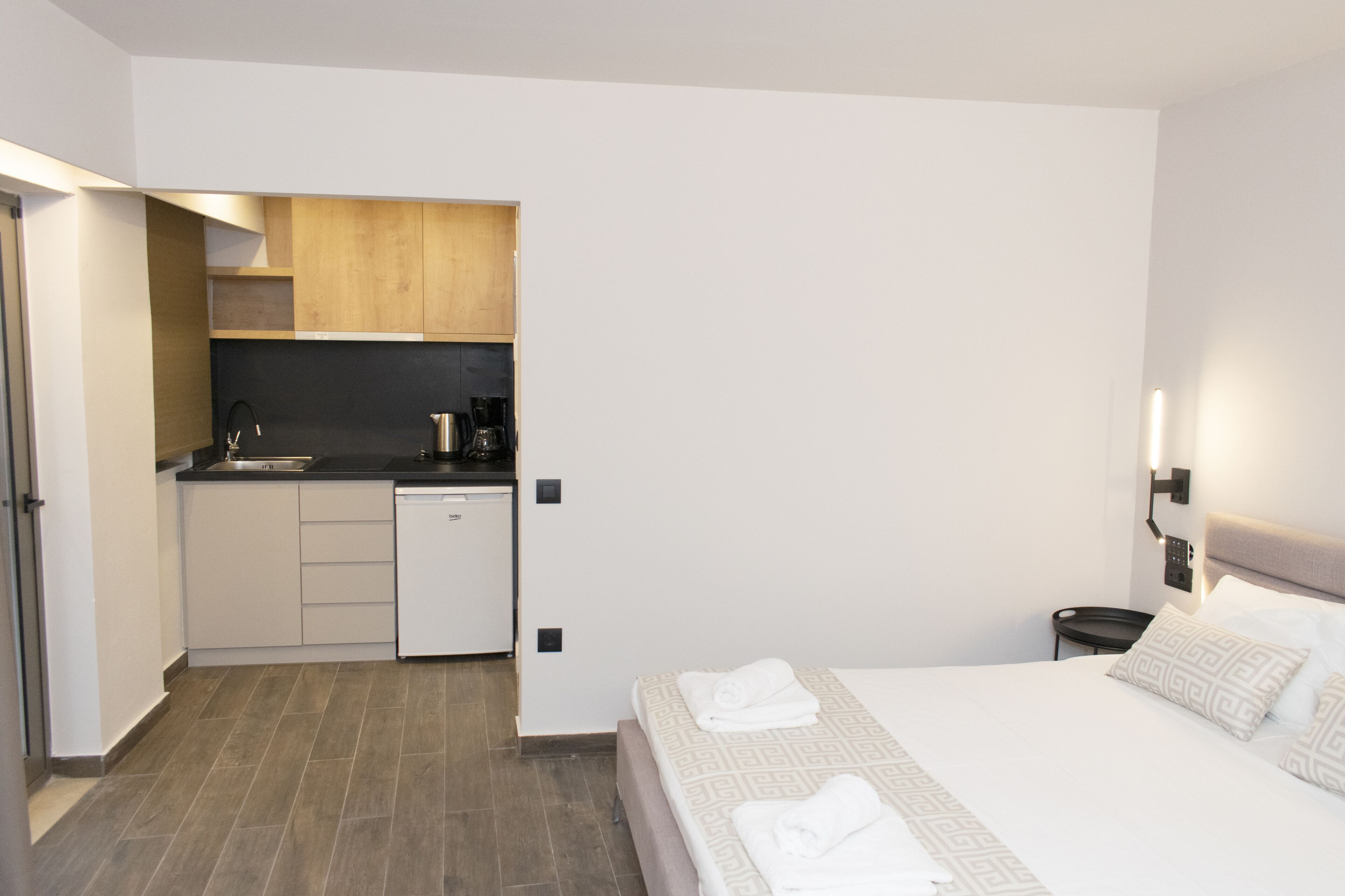 A modern hotel room with a separate kitchenette, a neatly made bed, and a small bedside table.
