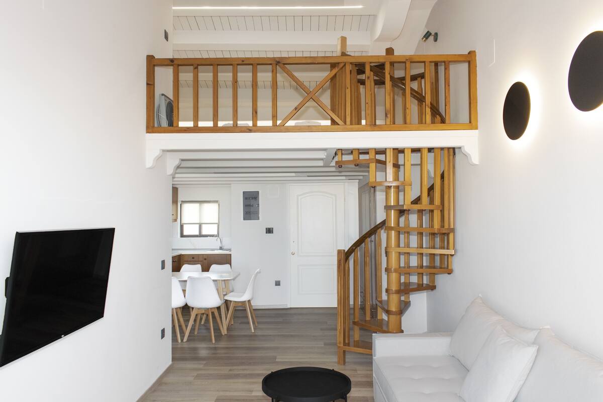 A spacious living area with a high ceiling, a wooden staircase, a flat-screen TV mounted on the wall, a white sofa, and a dining area with white chairs.