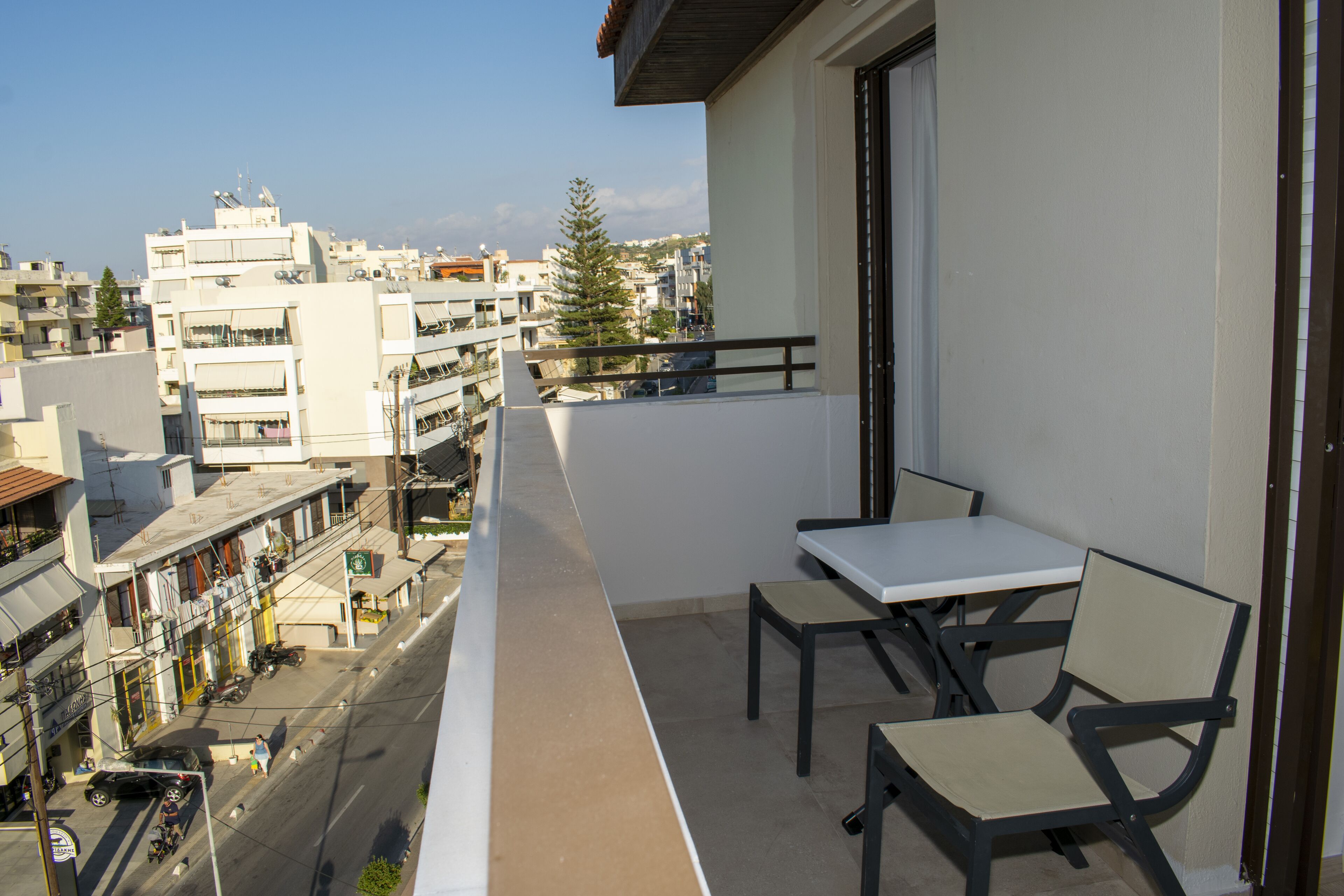 A balcony with a table and chairs, overlooking a street with buildings and a clear sky.