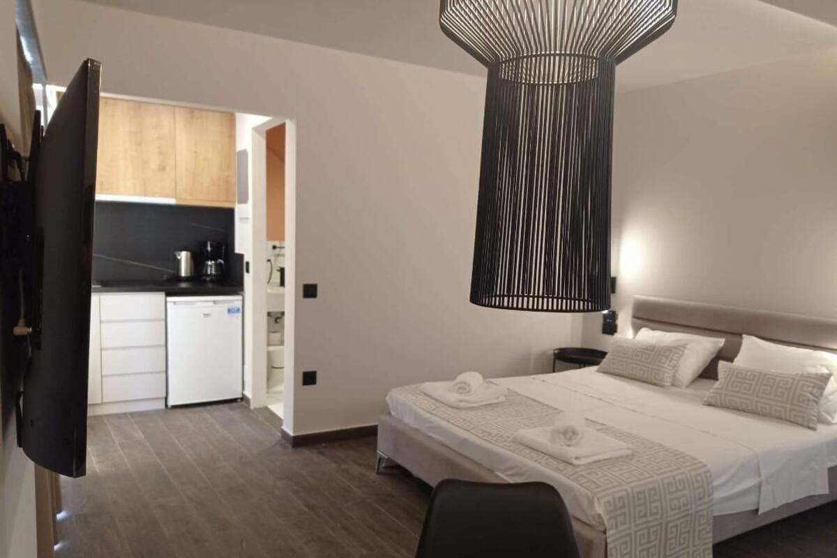 A modern hotel room with a large bed, a bedside table, a hanging light fixture, and a view of the kitchen area.