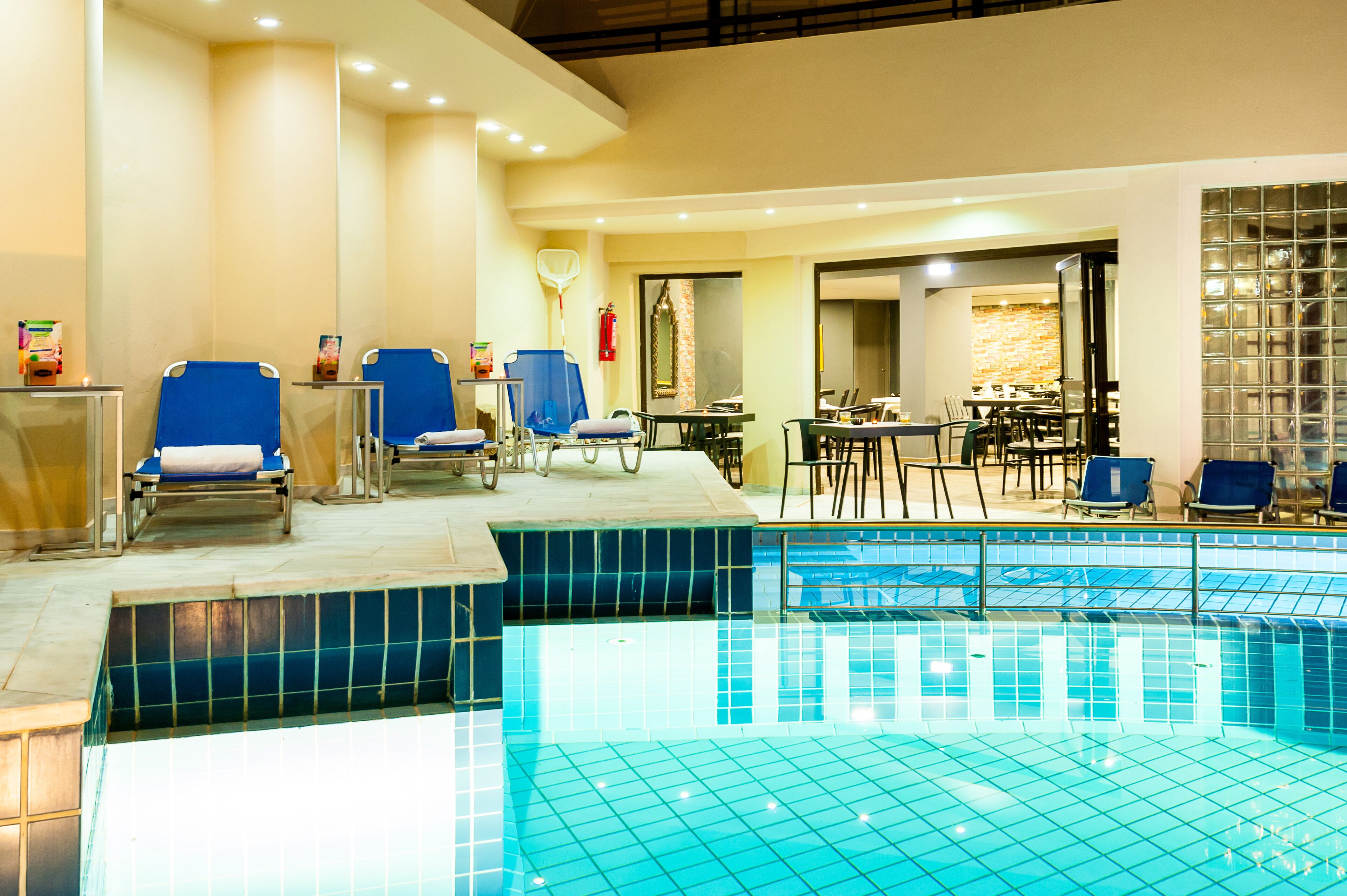 Une piscine intérieure aux murs recouverts de carreaux bleus, avec des chaises longues et un espace repas comprenant des tables et des chaises.