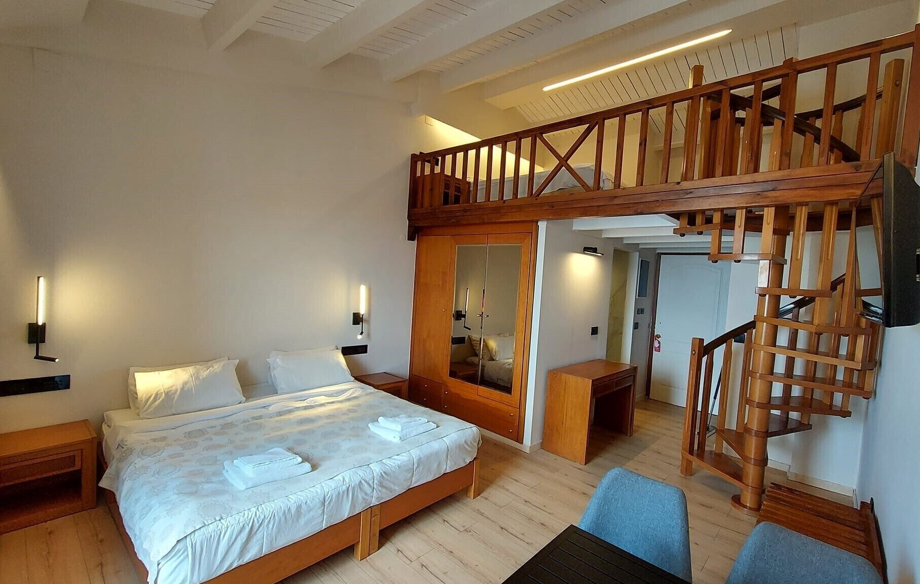 A bedroom with a bed, bedside tables, a mirror, a wooden staircase, and a small seating area.