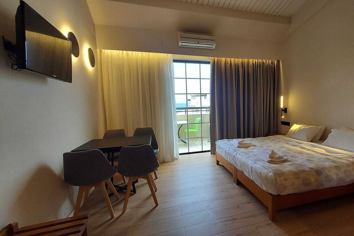 A hotel room with a bed, a desk with chairs, a TV, and a balcony with curtains.