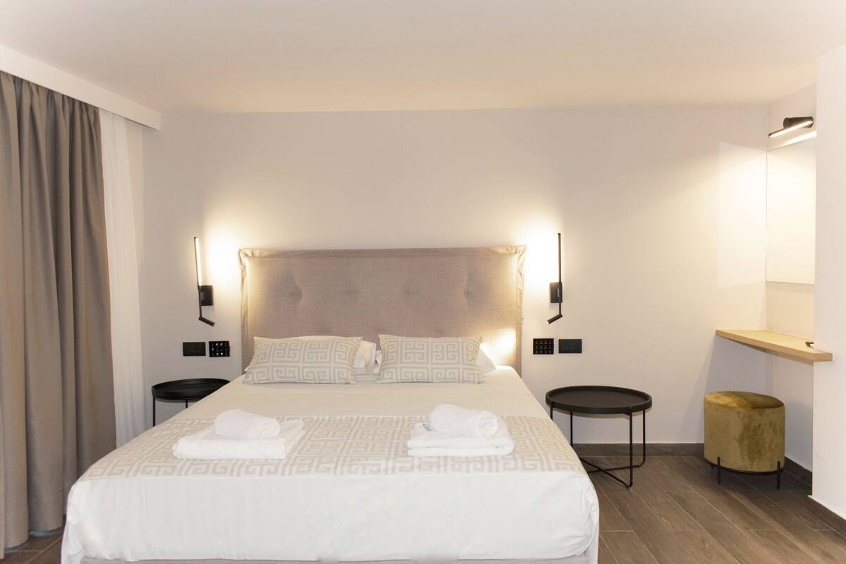 A modern hotel room with a large bed, two bedside tables, a round side table, a small stool, and a bedside lamp.