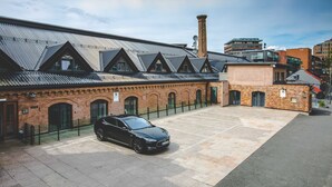 Un área de estacionamiento con un coche negro, un edificio de ladrillo con techo metálico y un edificio moderno al fondo.