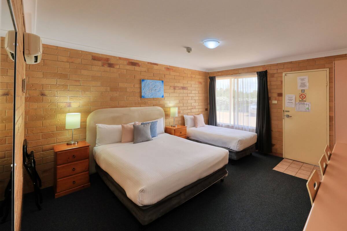 A hotel room with two beds, a brick wall, a window with curtains, a door with a sign, and a TV on the wall.
