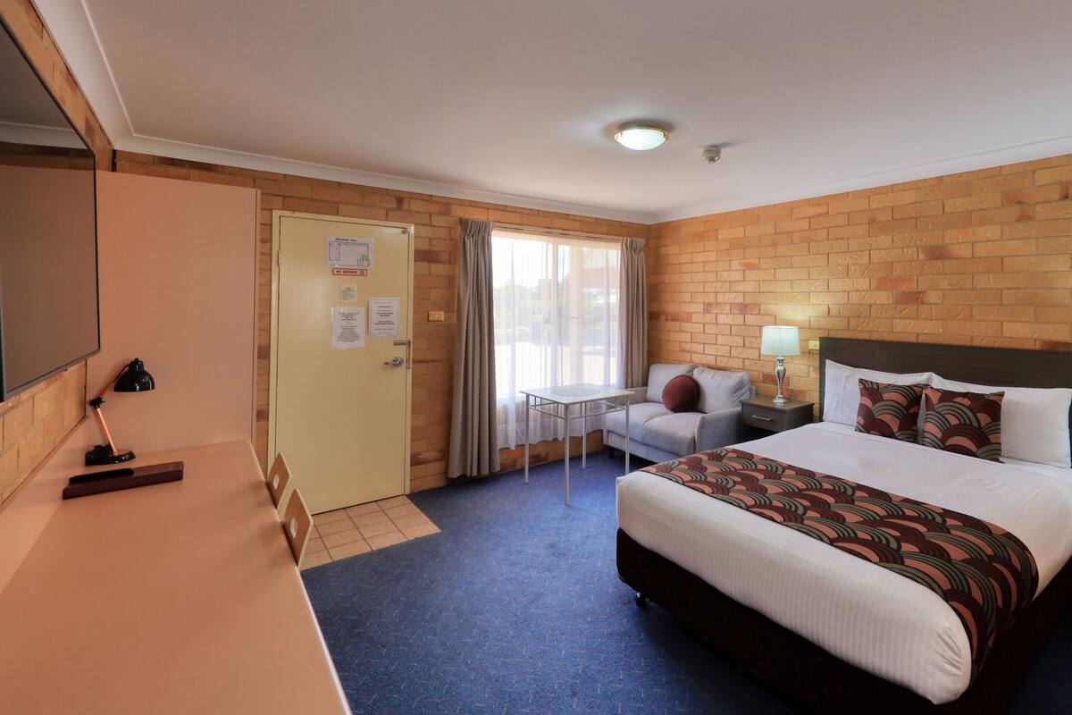 A hotel room with a bed, a desk, a chair, a nightstand, and a window with curtains.