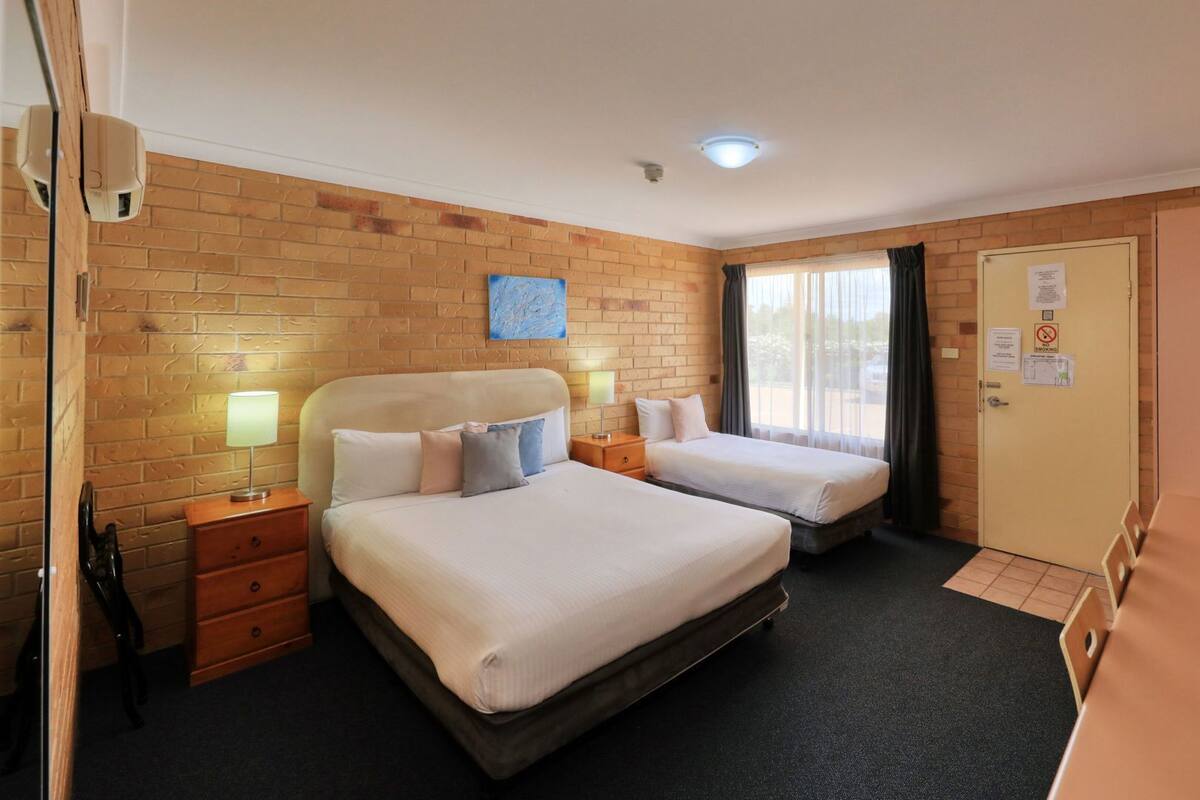 A hotel room with two beds, a brick wall, a window with curtains, a door with a no-smoking sign, and a television.