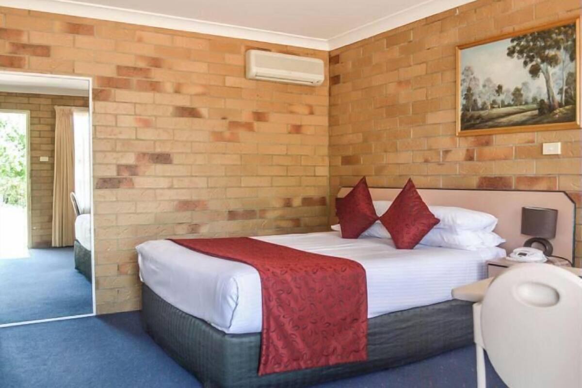 A hotel room with a brick wall, a bed with white linens and red pillows, a painting, and a bedside table with a lamp.