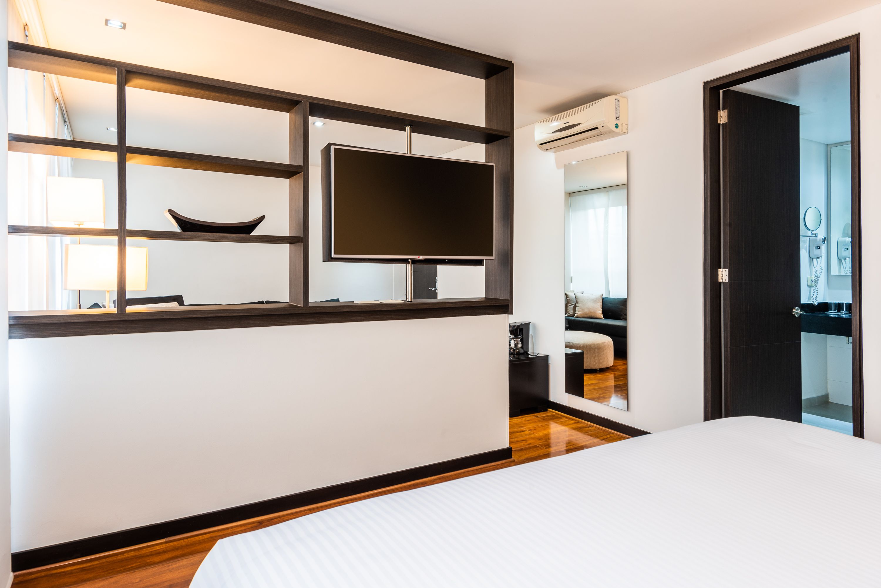 A modern hotel room with a large bed, a flat-screen TV mounted on a wooden shelf, a small table with a lamp, and a bathroom visible through a glass door.