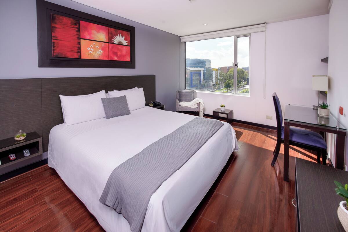 A modern hotel room with a large bed, a desk, a chair, a nightstand, and a window with a city view.