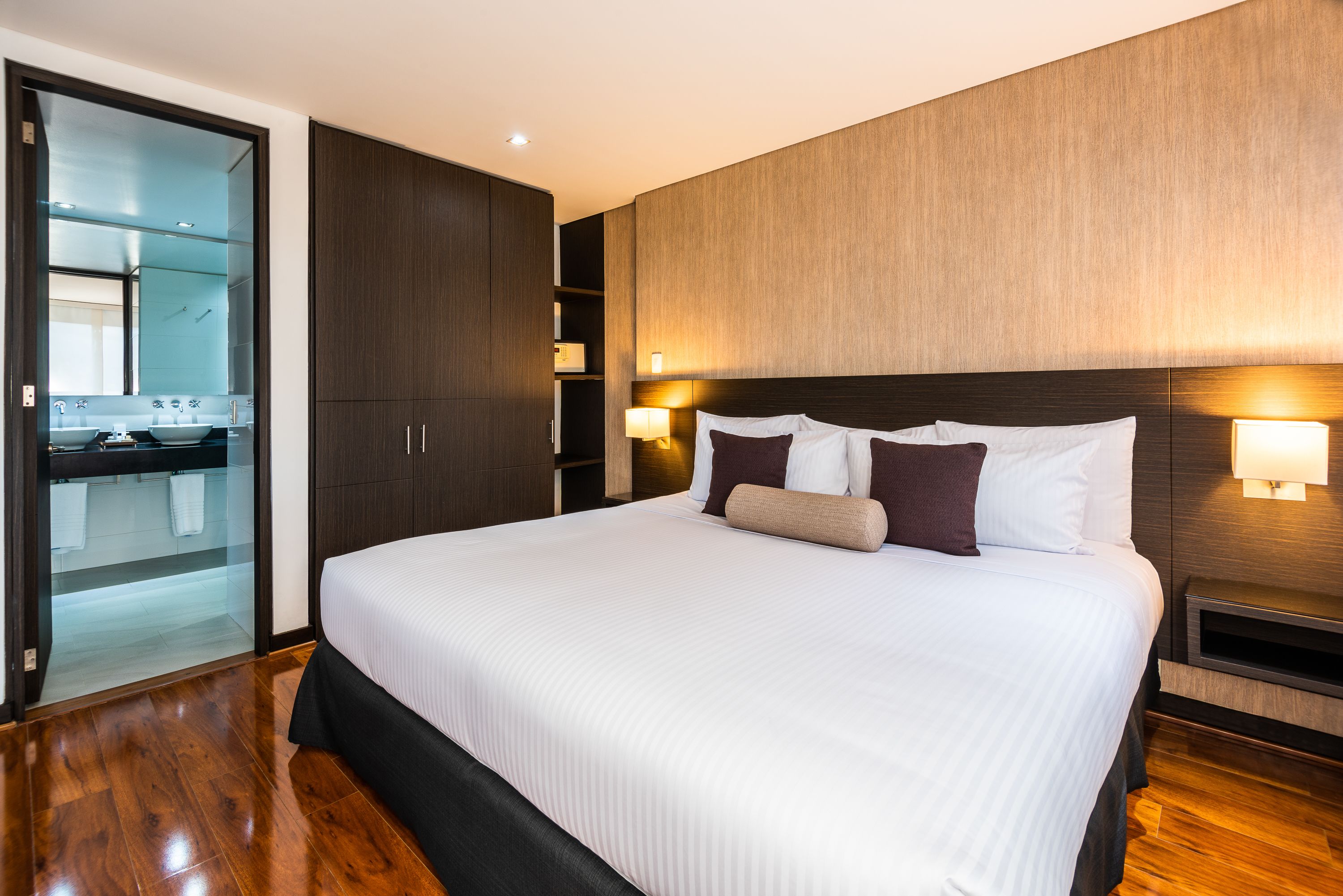 A modern hotel room with a large bed, wooden flooring, a built-in wardrobe, and a glass-enclosed bathroom.
