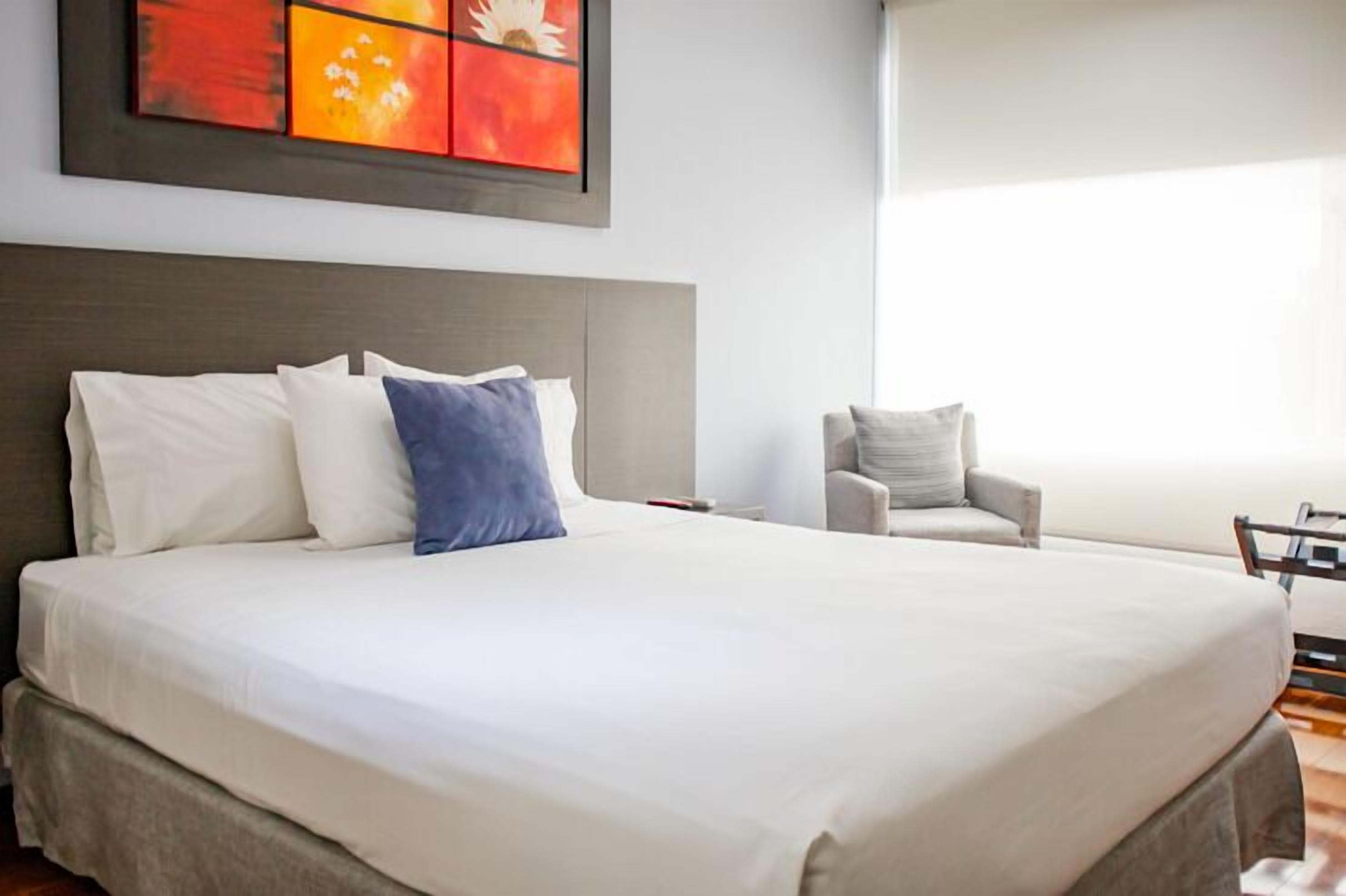 A large bed with white and blue pillows, a grey headboard, and a chair in a well-lit room with a large window.