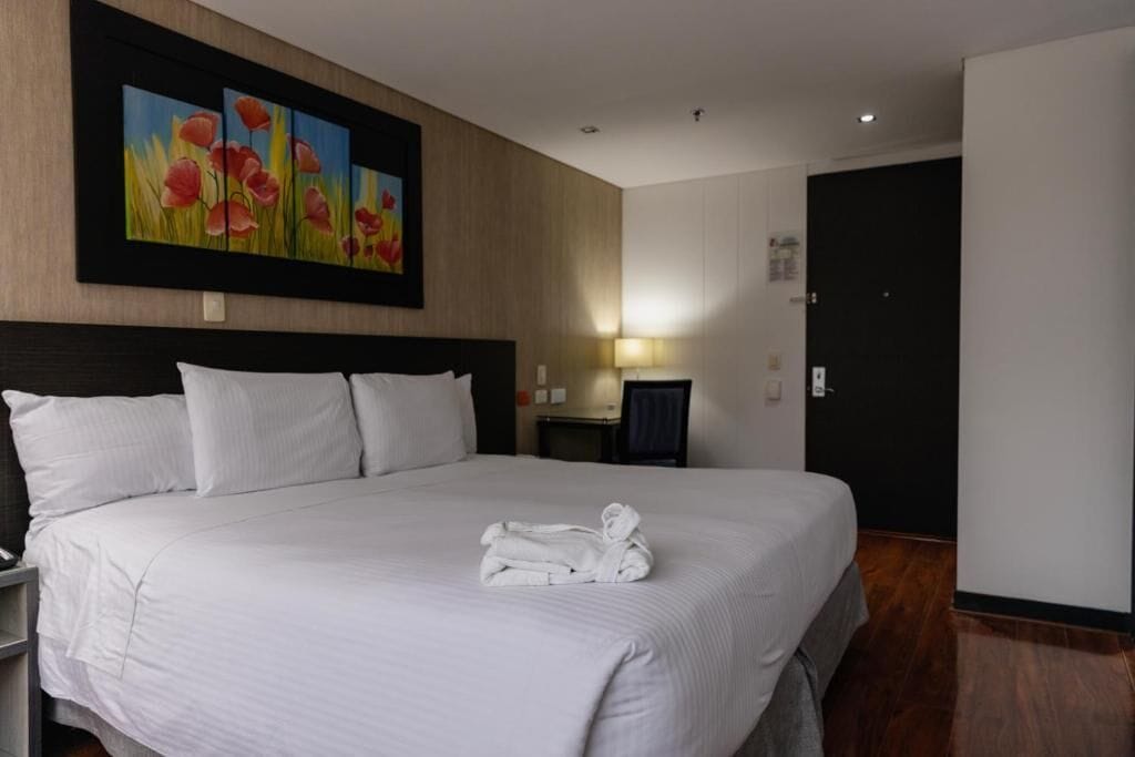 A hotel room with a large bed, white bedding, a painting of flowers on the wall, a bedside table with a lamp, and a door to the right.