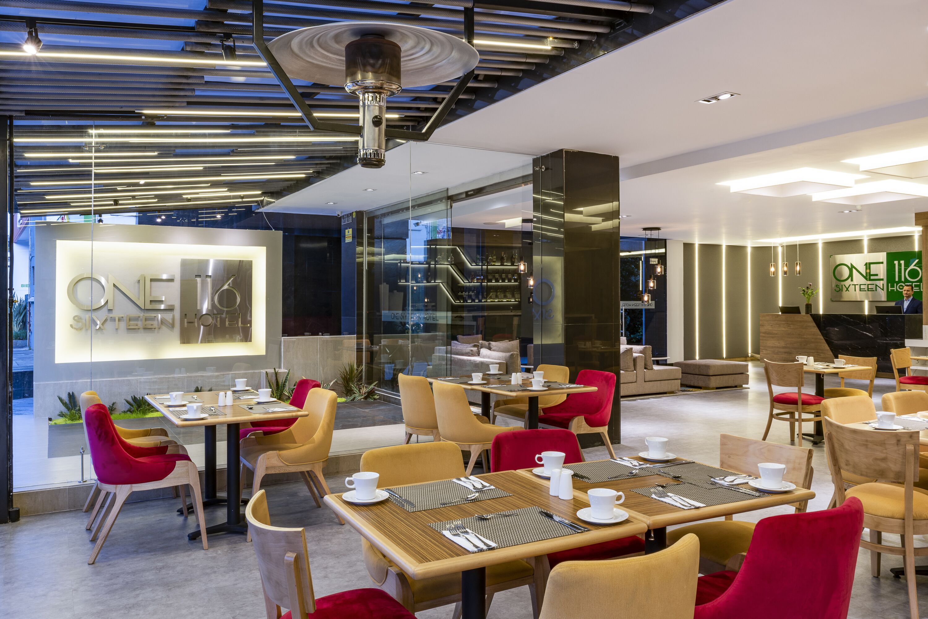A modern hotel lobby with a glass facade, wooden tables, and red and yellow chairs.