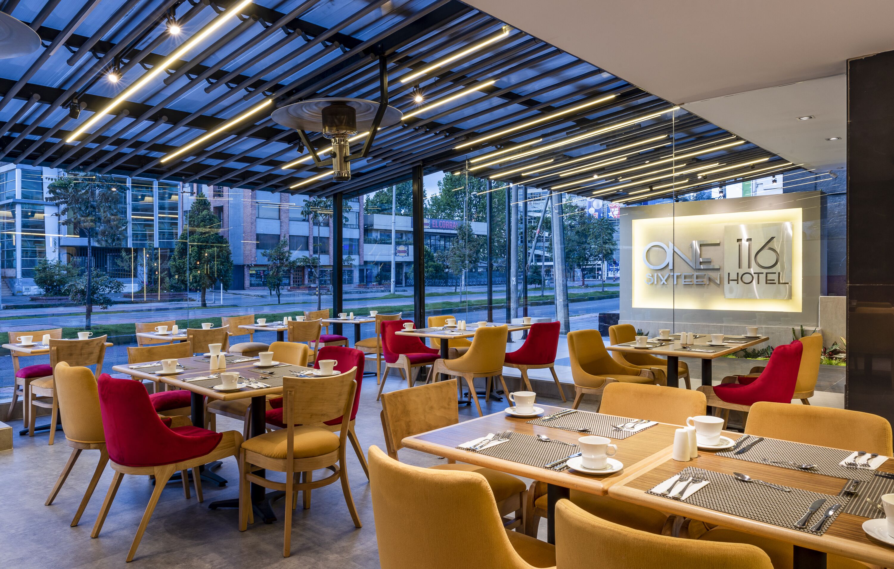 A modern hotel dining area with tables and chairs, a view of the street, and a sign for the hotel.
