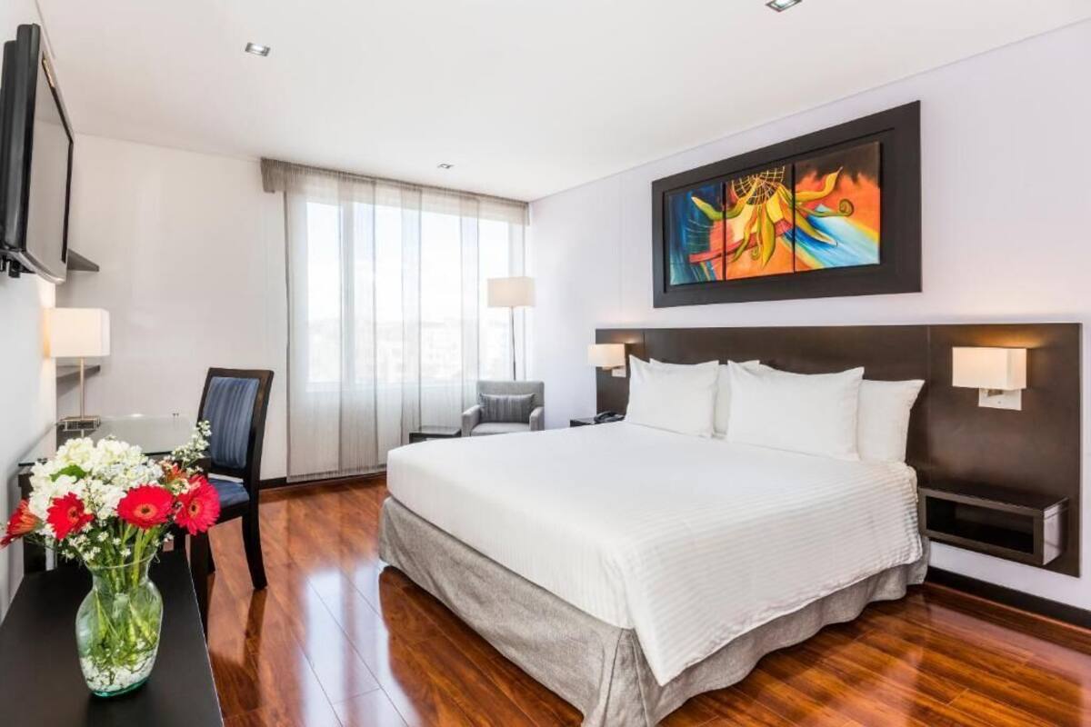 A hotel room with a large bed, a desk with a vase of flowers, a chair, a TV, and a painting on the wall.