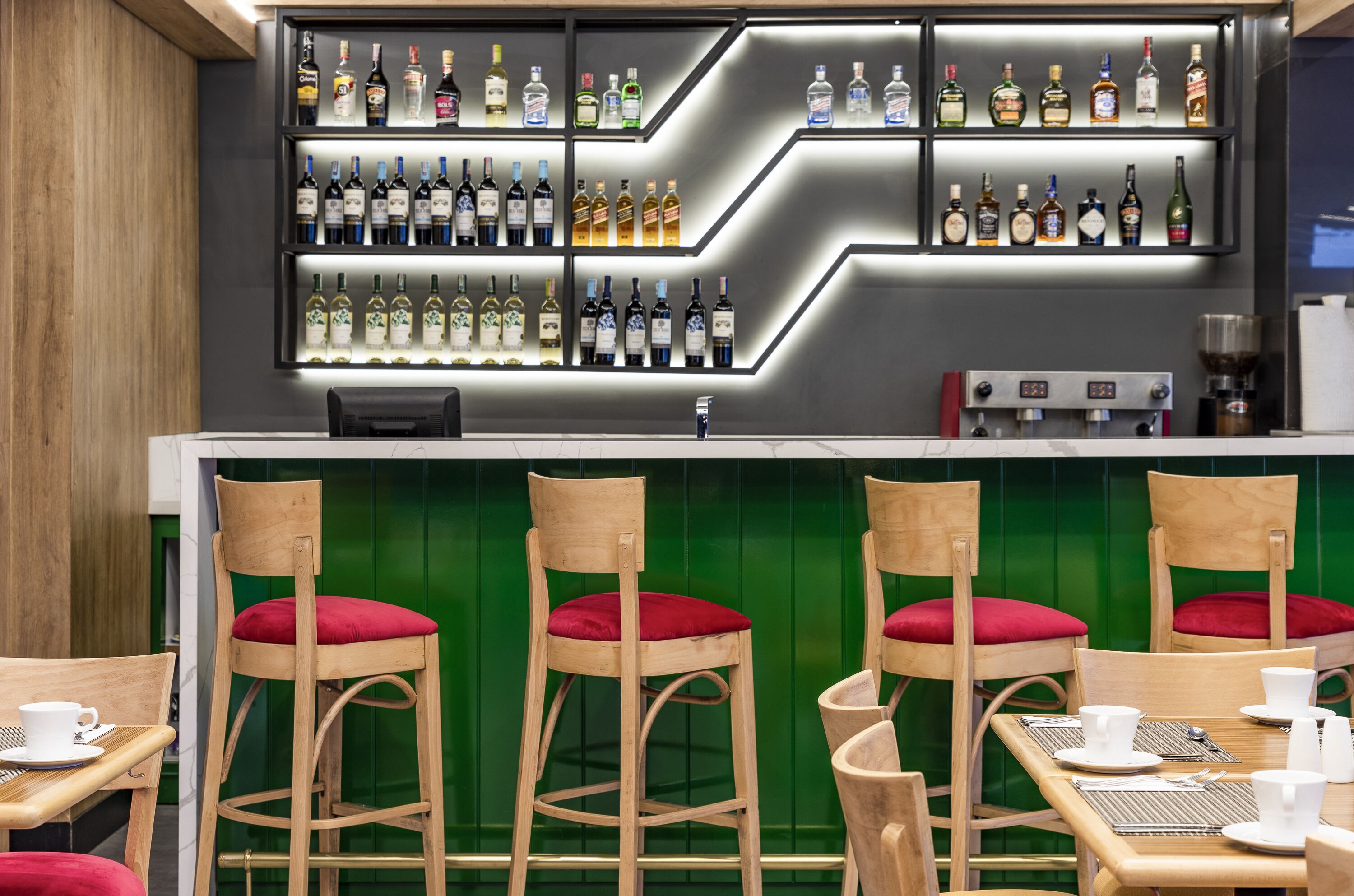 A bar area with a variety of liquor bottles, wooden bar stools with red cushions, and a coffee machine.