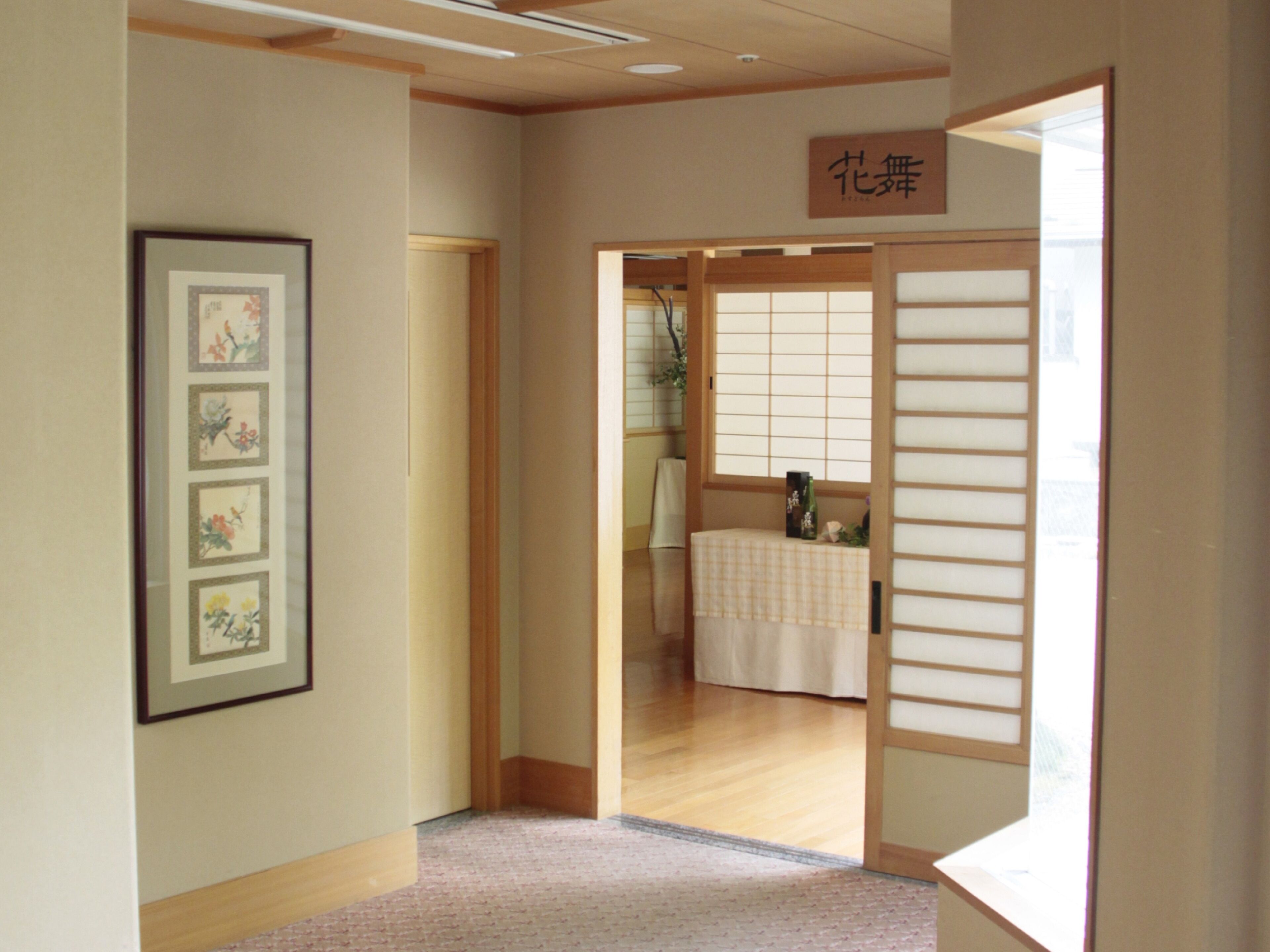 A hallway with a wooden floor, a framed picture on the wall, and a sliding door leading to a room with a tatami mat and a small table.