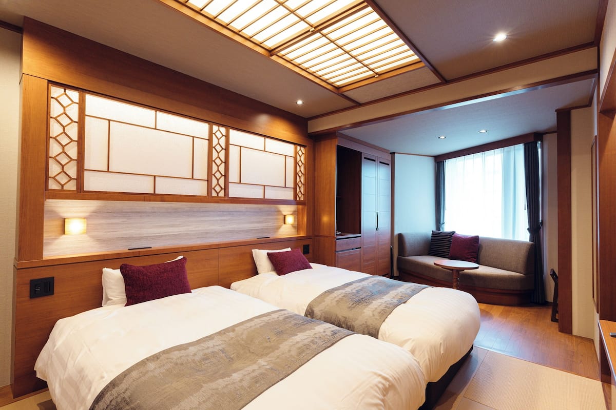 A hotel room with two beds, a sofa, a small table, and a view of the outside.