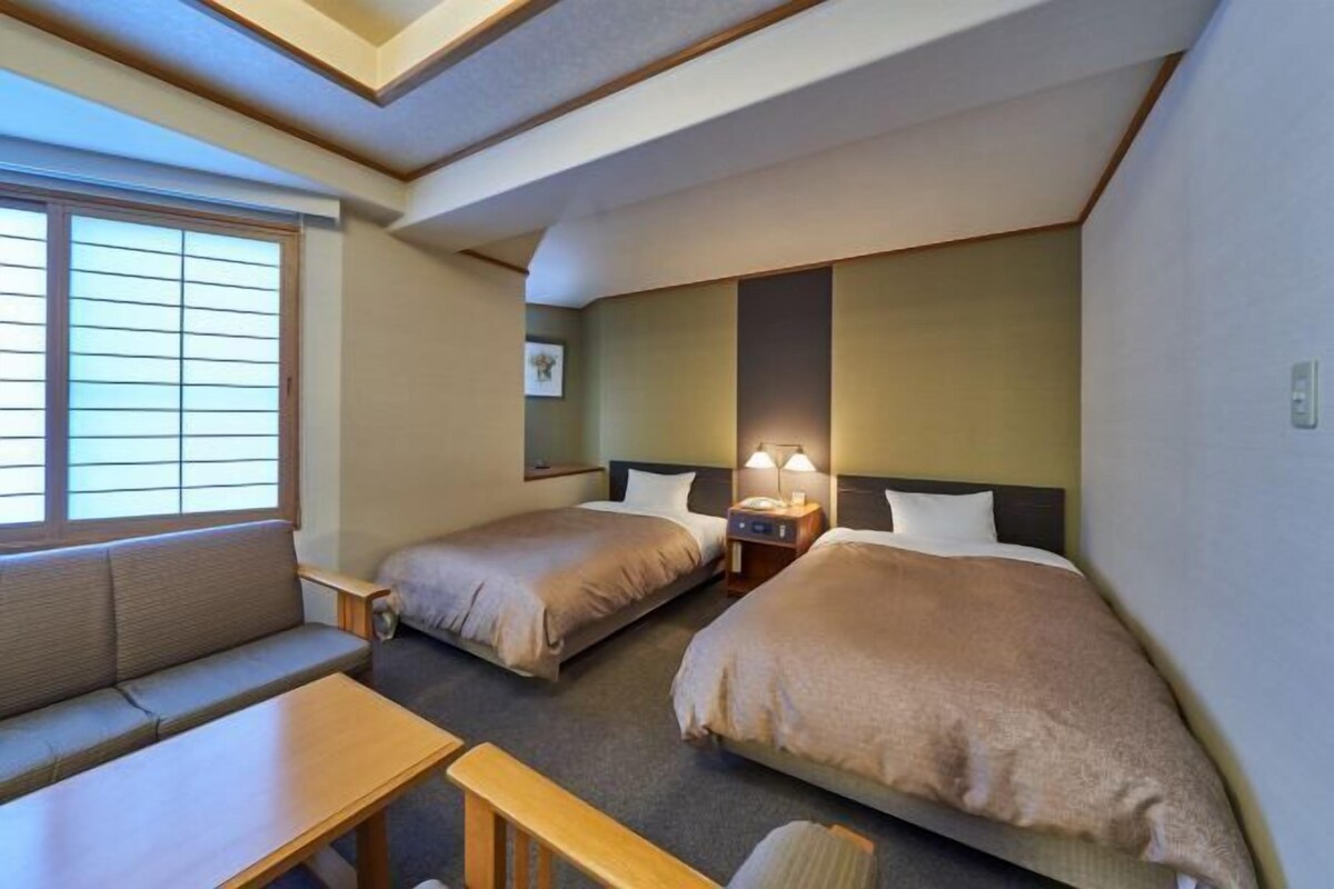 A hotel room with two beds, a sofa, a table, and a window with blinds.