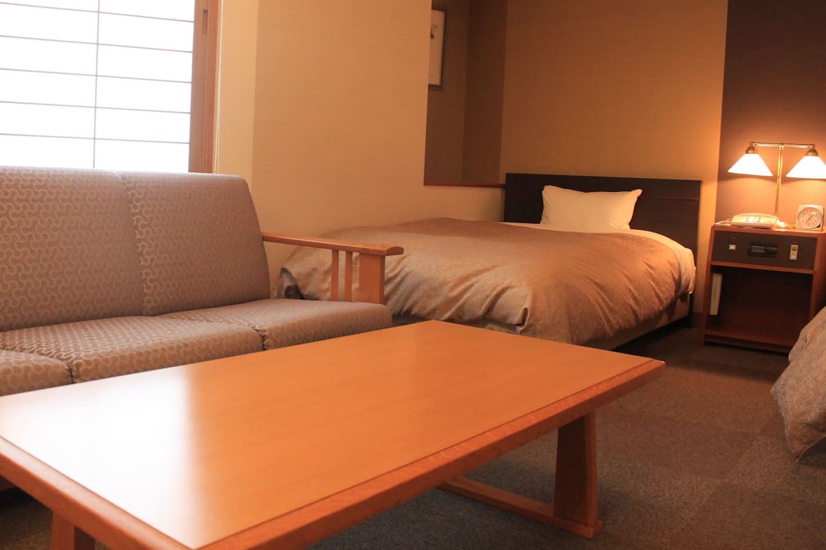 A hotel room with a sofa, a single bed, a nightstand with a lamp, and a small table.