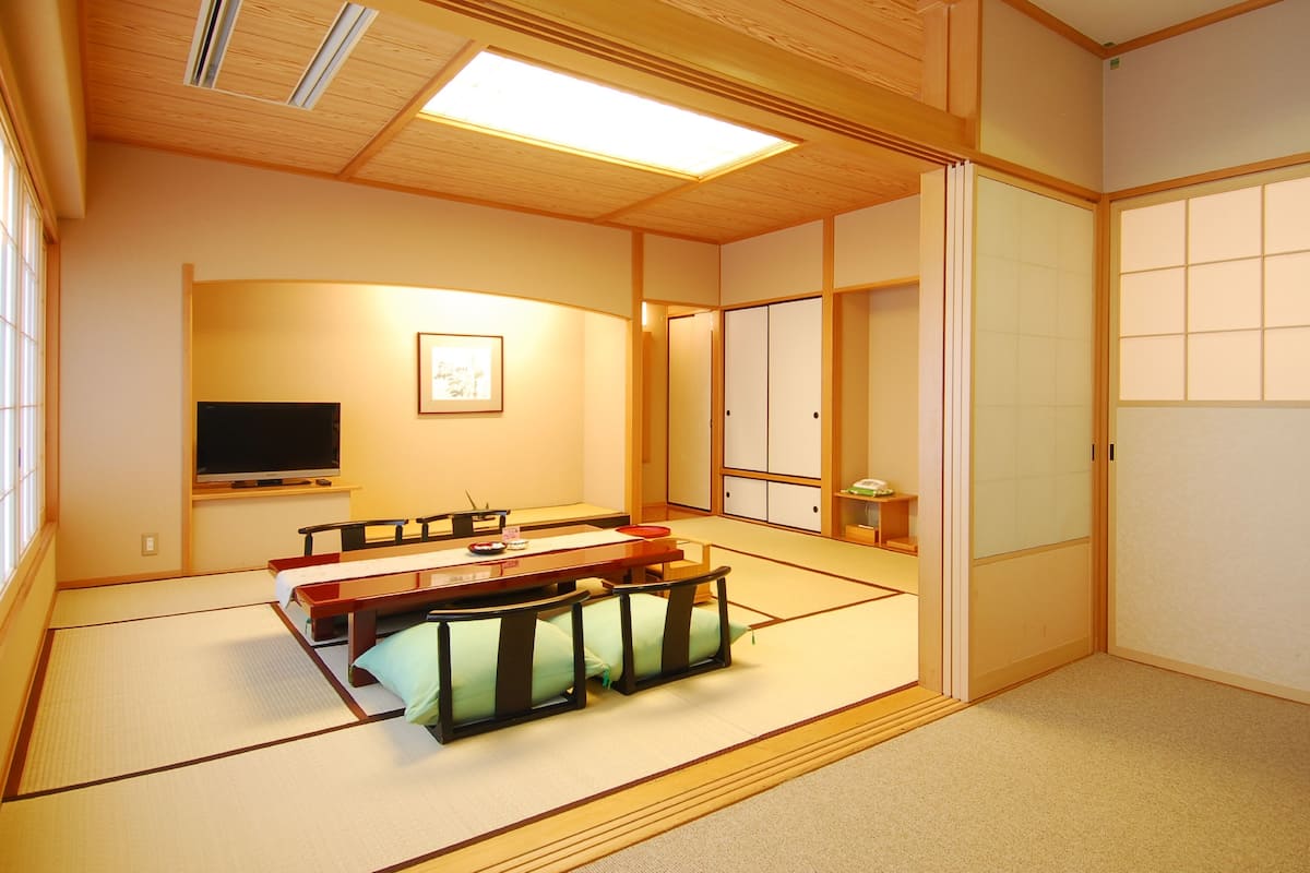 A traditional Japanese room with tatami flooring, a low table with cushions, a television, sliding doors, and a large window.