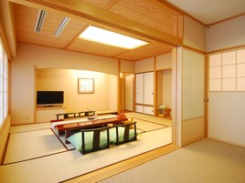 Una habitación japonesa tradicional con suelo de tatami, una mesa baja con cojines, televisión, puertas correderas y un ventanal grande.