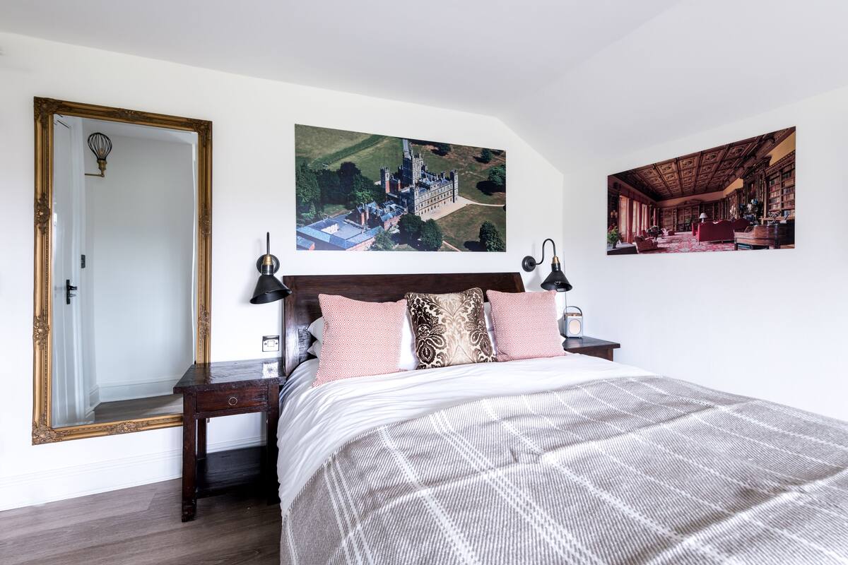Quarto com uma cama grande, uma mesinha de cabeceira de madeira, um espelho e duas fotos emolduradas na parede.