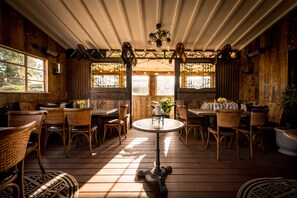 A rustic wooden interior with round tables and wicker chairs, large windows with plants, and a chandelier. - Queens Arms (Hungerford)