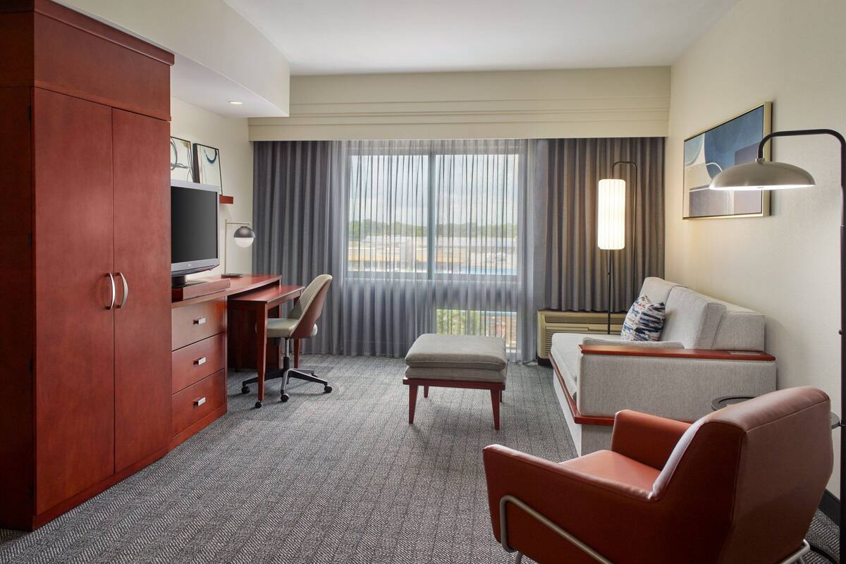 Habitación de hotel con escritorio, sofá, sillón rojo, televisor y ventana con cortinas.